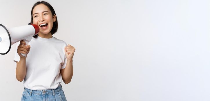 Asian girl shouting at megaphone, young activist protesting, using loud speakerphone, making announcement, white background. Copy space