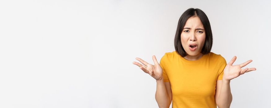 Frustrated, confused asian woman cant understand smth, looking clueless, shrugging shoulders, standing in yellow tshirt over white background.