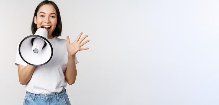 Happy asian woman shouting at megaphone, making announcement, advertising something, standing over white background.