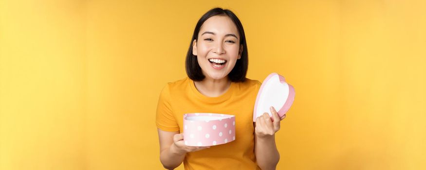 Portrait of excited asian woman, open gift box with surprised happy face, standing over yellow background.