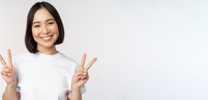 Cute asian girl showing peace, v-sign, smiling and looking happy at camera, wearing white tshirt, studio background.