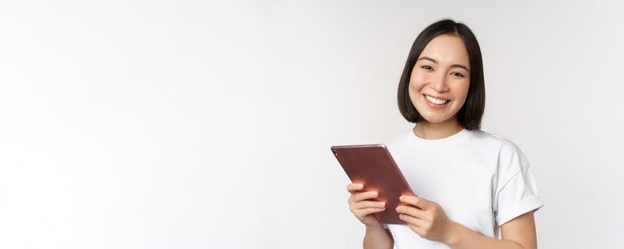 Smiling asian girl with digital tablet, looking happy and laughing, posing in tshirt over white background. Copy space