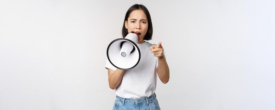 Angry asian woman with megaphone, scolding, accusing someone, protesting with speakerphone on protest, standing over white background.