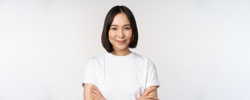 Close up portrait of confident korean girl, student looking at camera with pleased smile, arms crossed on chest, standing over white background.