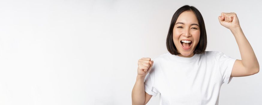 Portrait of enthusiastic asian woman winning, celebrating and triumphing, raising hands up, achieve goal or success, standing over white background.