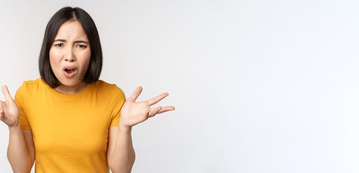 Frustrated, confused asian woman cant understand smth, looking clueless, shrugging shoulders, standing in yellow tshirt over white background.