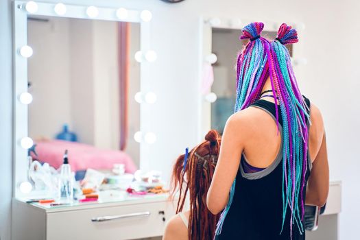 Back view of female barber with colored pigtails, who braids ginger dreadlocks to woman. Creative and fashionable hairstyle. Service in beauty salon. Large mirror with light bulbs on background.