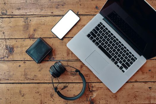 Laptop, wireless headphones, mobile phone, and wallet on wooden table. Freelancer's work surface. Blank cell phone screen to insert some text. Modern devices for business jobs and entertainment.