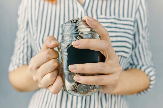 Glass bottle with coins in women's hands. Blank black sticker on donation money jar. Girl in white and black striped shirt holds her savings. Pot for tips.