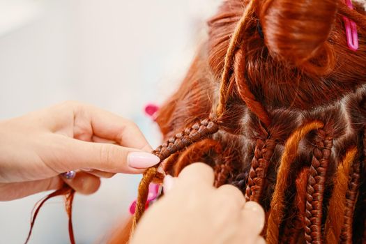 Hairdresser weaves woman ginger dreadlocks. Bright pigtails on head. Hippie style hairstyle. Beauty salon services. Close up of braiding process african plaits with colored kanekalon.