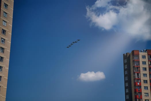 Group of Russian air force demonstrates aerobatics. Military aviation victory parade over city multi-storey buildings. Presentation of aviation equipment. White clouds and rays of summer sun in sky.