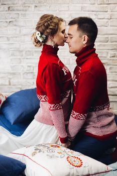 Side view of romantic couple in similar red Xmas sweater kissing holding hands surrounded by pillows against white brickwall.