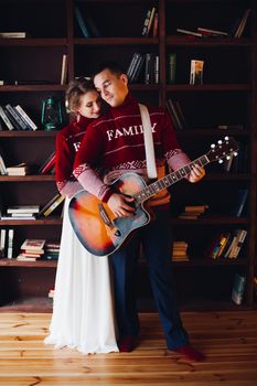 Couple wearing in red sweaters, sitting on floor at library and playing on gitar, and embracing. Happy family romantic spending time together. Winter holidays, x mas, new year concept.