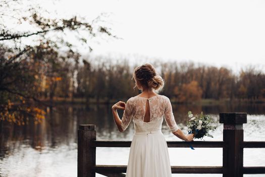 Back view of blonde bride in beautiful white dress with bouquet in hand standing against lake in forest on autumn day.