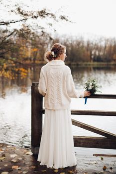 Back view of beautiful blonde bride standing at river pear, leaning on wooden fence and looking away at distance. Stylish woman in wedding dress and sweater posing alone. Conceptual autumn wedding.