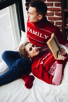 Family of young pretty blonde woman lying on knees of her boyfriend, who looking at window. Couple in red sweaters relaxing, spending tome together. Winter holiday. Christmas look.