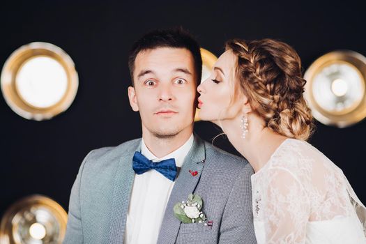 Portrait of handsome elegant fiance with blue bow-tie looking at camera with open wide eyes while bride kissing him in cheek. Big round lights in background.