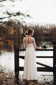 Back view of blonde bride in beautiful white dress with bouquet in hand standing against lake in forest on autumn day.