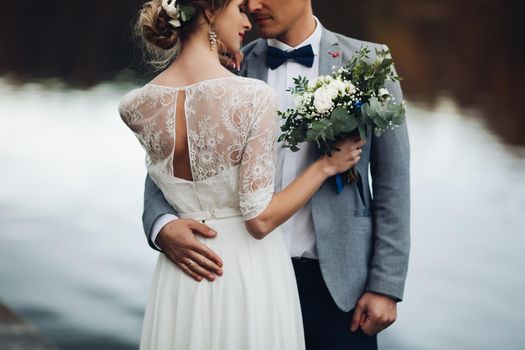 Back view of husband embracing his beautiful elegant wife in lace wedding dress with lovely bouquet against unfocused lake surface.