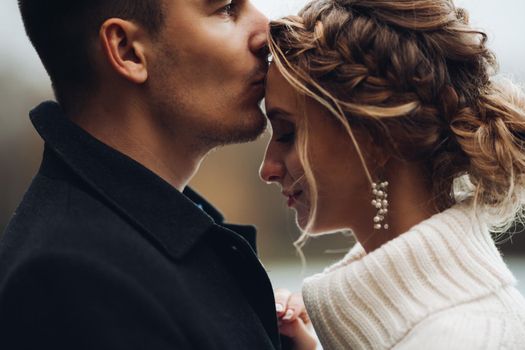 Close up of young beautiful couple of blonde bride and handsome groom posing outdoors at cold weather and kissing. Husband kissing beautiful wife in cheen, she smiling. Autumn wedding.