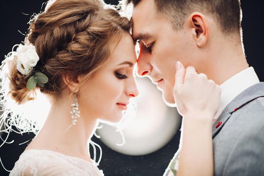 Portrait of beautiful young couple young gorgeous bride and handsome groom at rounded light background. Sweet, beloved sweathearts leaning by nose each other, embracing and touching by faces. Wedding.