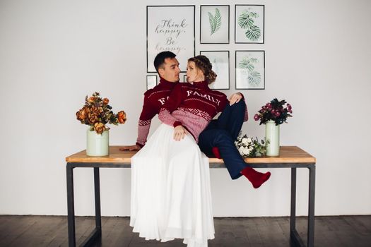 Full length portrait of loving newlymarried couple sittng on table and looking face to face. Both wearing similar red Xmas sweater with Family word against modern posters and vases of flowers on table.