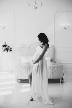 View from side of future mother in beautiful white dress standing in bedroom, looking at camera, smiling and posing. Adorable pregnant woman expecting little child. Concept of pregnancy and love.