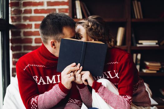 Portrait of loving couple in similar red knitted sweater with family word on them kissing hiding behind the book at home.