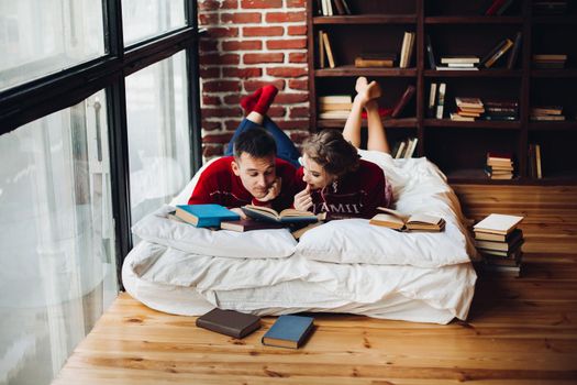 Portrait of romantic couple in similar red sweater lying on mattress reading different books together at home. Christmas time.