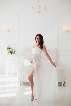 View from side of future mother in beautiful white dress standing in bedroom, looking at camera, smiling and posing. Adorable pregnant woman expecting little child. Concept of pregnancy and love.