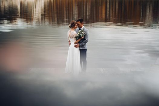 Panoramic view at youg couple bride in whire and groom in gray standing among lake, posing and embracing. Conceptual wedding at nature outdoor in autumn swasone. Wedding, family, love story concept.
