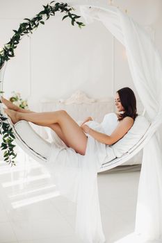 Full length portrait of stunning pregnant brunette woman in elegant white dress with transparent skirt sitting on modern suspended chair in beautifully decorated white bedroom. She is looking at camera.