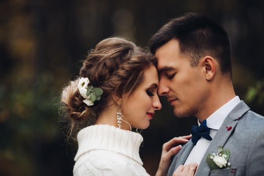 Beautiful couple of bride and groom celebrating wedding at autumn season. Gorgeous wife in white and handsome groom in gray suite touching by face with clossed eyes, smiling, embracing outdoors.