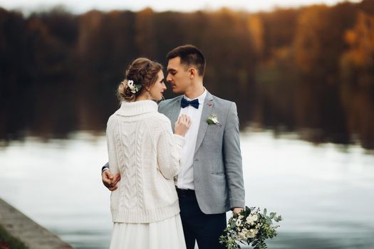 Back view of husband embracing his beautiful elegant wife in lace wedding dress with lovely bouquet against unfocused lake surface.