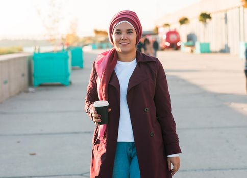 Young Arabic beautiful woman in hijab walking at street, tapping on smartphone and drinking coffee to-go. Pretty muslim female in headscarf strolling at aeroport, texting message and sipping drink.