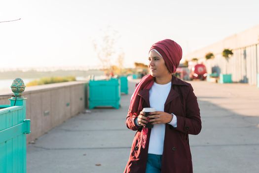 Young Arabic beautiful woman in hijab walking at street, tapping on smartphone and drinking coffee to-go. Pretty muslim female in headscarf strolling at aeroport, texting message and sipping drink.