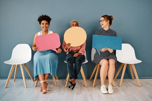 Studio shot of a group of attractive young businesswomen holding speech bubbles while sitting in a row against a grey background.