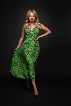 Front view of slim blonde standing on black isolated background and posing. Attractive young woman in green long dress looking at camera and smiling in studio. Concept of elegance and glamour.