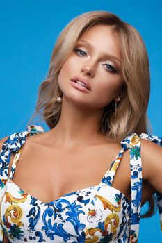 Portrait of sexy young blonde with makeup and hairstyle looking at camera and posing in studio. Beautiful woman wearing accessories standing on blue isolated background. Concept of beauty.