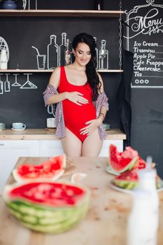 Focus on cut juicy watermelon on wooden table. Unfocused blurred background with unrecognizable pregnant brunette woman in red embracing her belly.