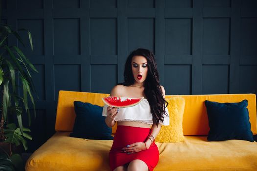 Stylish pregnant woman sitting on orange sofa, eating watermelon and posing. Attractive future mother in elegant clothes awaiting for little baby and relaxing. Concept of pregnancy and fashion.
