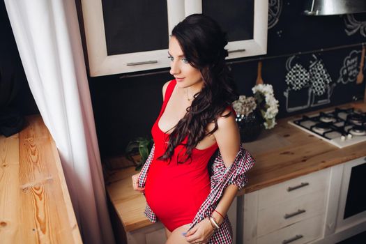 Adorable pregnant woman standing at kitchen and touching big belly with hands. Attractive brunette expecting little child and enjoying motherhood. Concept of love and pregnancy.