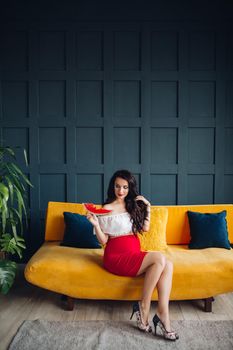 Stylish pregnant woman sitting on orange sofa, eating watermelon and posing. Attractive future mother in elegant clothes awaiting for little baby and relaxing. Concept of pregnancy and fashion.