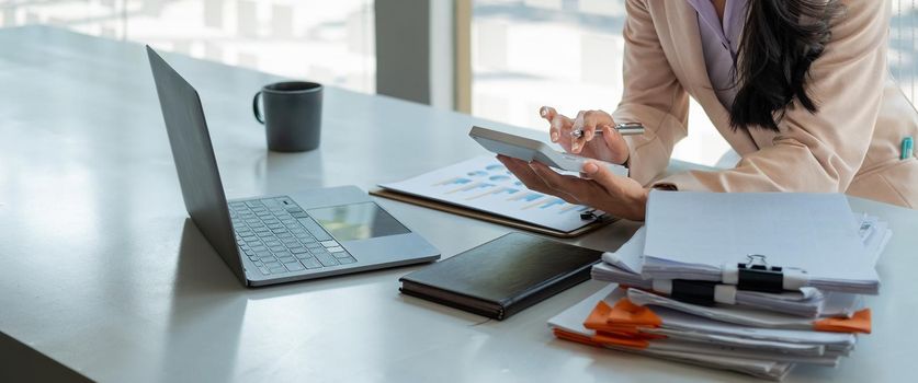 Close up of businesswoman or accountant hand holding pen working on calculator to calculate business data, accountancy document and laptop computer at office, business concept.