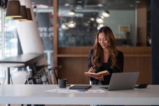 Happy asian business woman working on laptop computer and taking notes at the office.