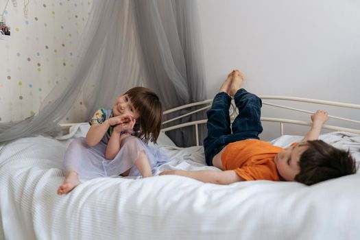 Two siblings happily playing together on the sofa bed covered with white blanket in the kids' room smiling into the camera. High quality photo