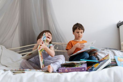 Two siblings reading books together on the sofa bed covered with white blanket in the kids' room, one of them laughing into the camera