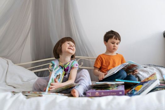 Two siblings reading books together on the sofa bed covered with white blanket in the kids' room, one of them laughing into the camera