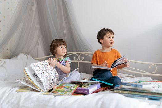 Two siblings reading books together on the sofa bed covered with white blanket in the kids' room, while both of them are communicating with each other