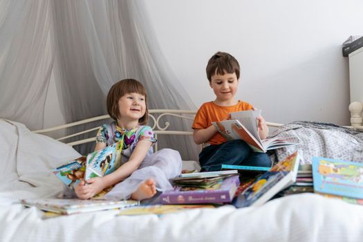 Two siblings reading books together on the sofa bed covered with white blanket in the kids' room, while both of them are smiling